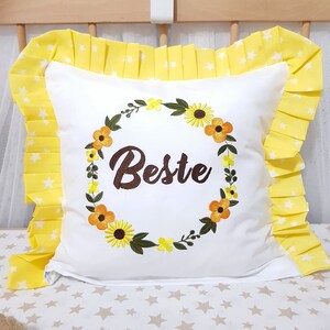 May include: White pillow with a yellow ruffle border and a floral wreath embroidered on the front. The wreath is made of green leaves and yellow and orange flowers. The word "Beste" is embroidered in brown inside the wreath.