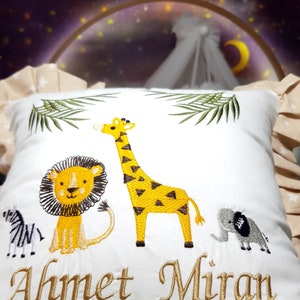 Könnte beinhalten: Ein weißes Kissen mit gestickten Bildern einer Giraffe, eines Löwen, eines Zebras und eines Elefanten. Das Kissen trägt den Namen "Ahmet Miran", der in goldener Farbe gestickt ist.