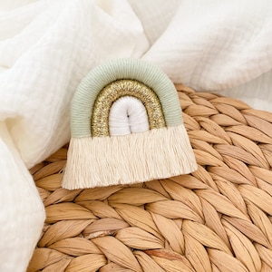 Peut inclure: Un petit arc-en-ciel fait main en laine avec un schéma de couleurs vert, or et blanc. L'arc-en-ciel a une frange en bas.