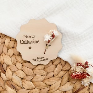 Peut inclure: Un présentoir en bois clair aux bords festonnés, avec l'inscription "Merci Catherine" et un petit cœur. Un petit bouquet de fleurs séchées est attaché. Le texte "Emma MS 2024-2025" est également présent.