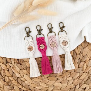 Op de afbeelding: Vier macramé sleutelhangers met houten accenten en kwastjes. De sleutelhangers hebben verschillende kleuren, waaronder wit, roze en fuchsia. Elke sleutelhanger heeft een houten hart met tekst, waaronder "Maman", "Yours" en "Home".
