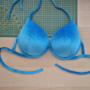 Könnte beinhalten: Ein blaues Samt-Bikinioberteil mit Neckholder-Trägern und Unterbrustbändern. Das Oberteil hat einen leuchtenden Blauton mit einer weichen, plüschigen Textur. Das Bikinioberteil ist für Bademode konzipiert.