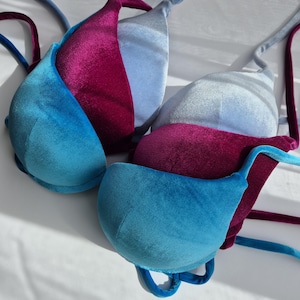 Puede incluir: Tres tops de bikini de terciopelo en diferentes colores: azul verdoso, azul claro y burdeos. Los tops están apilados uno encima del otro, con el top azul verdoso en la parte inferior y el top burdeos en la parte superior.