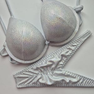 May include: A silver bikini set with a shimmering, textured finish. The set includes a padded, triangle-style top with adjustable straps and a matching thong-style bottom. The fabric has a subtle, iridescent quality.