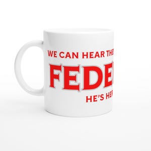 Puede incluir: Taza de cerámica blanca con asa curva. La taza presenta el texto rojo "WE CAN HEAR THE FED" y "HE'S HERE" en negrita, sin serifa. La taza es de tamaño estándar para café o té.