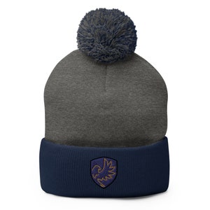 May include: A gray and navy blue beanie with a pom pom on top. The beanie has a navy blue cuff with an embroidered purple and gold bird logo.