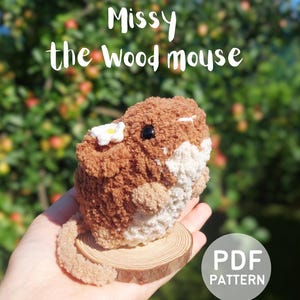 May include: A brown and white crocheted mouse plushie with a small white flower on its ear, sitting on a circular wooden slice. The text "Missy the Wood mouse" and "PDF PATTERN" are visible in the image.