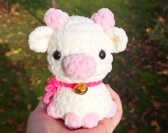 May include: A white crocheted cow with pink ears, nose, and hooves. The cow is wearing a pink ribbon around its neck and a gold bell.
