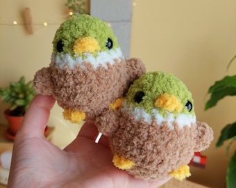 May include: Two small crocheted bird plushies with green heads, brown bodies, white accents, and yellow beaks. The plushies are held in a person's hand.