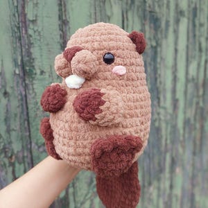 May include: A handmade, plush beaver toy in shades of brown. The stuffed animal has a rounded body, small ears, and a flat tail. It features dark brown paws, a white tooth, and a pink cheek. The toy is held up against a green background.
