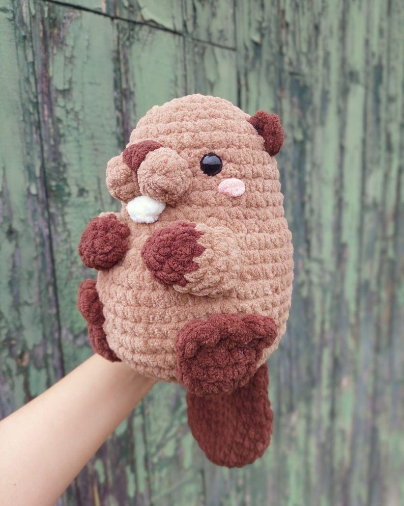May include: A handmade, plush beaver toy in shades of brown. The stuffed animal has a rounded body, small ears, and a flat tail. It features dark brown paws, a white tooth, and a pink cheek. The toy is held up against a green background.