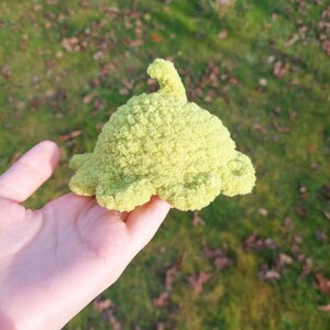 May include: A green crocheted plush toy shaped like a whale with a fin on top. The toy is being held in a person's hand.