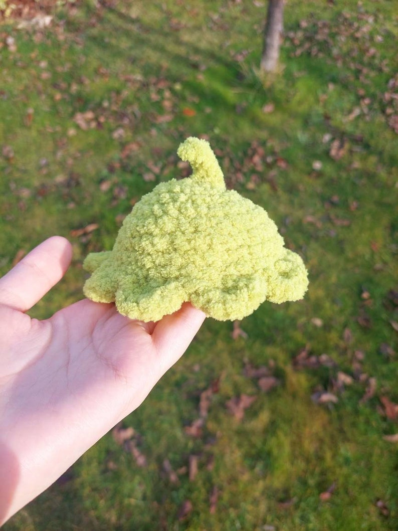 May include: A green crocheted plush toy shaped like a whale with a fin on top. The toy is being held in a person's hand.