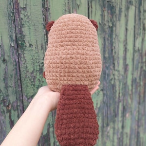 May include: A handmade, plush beaver toy in shades of brown. The body is a light brown, with darker brown ears and a large, flat tail. The toy is made of a soft, textured material, and is designed to be cuddly.