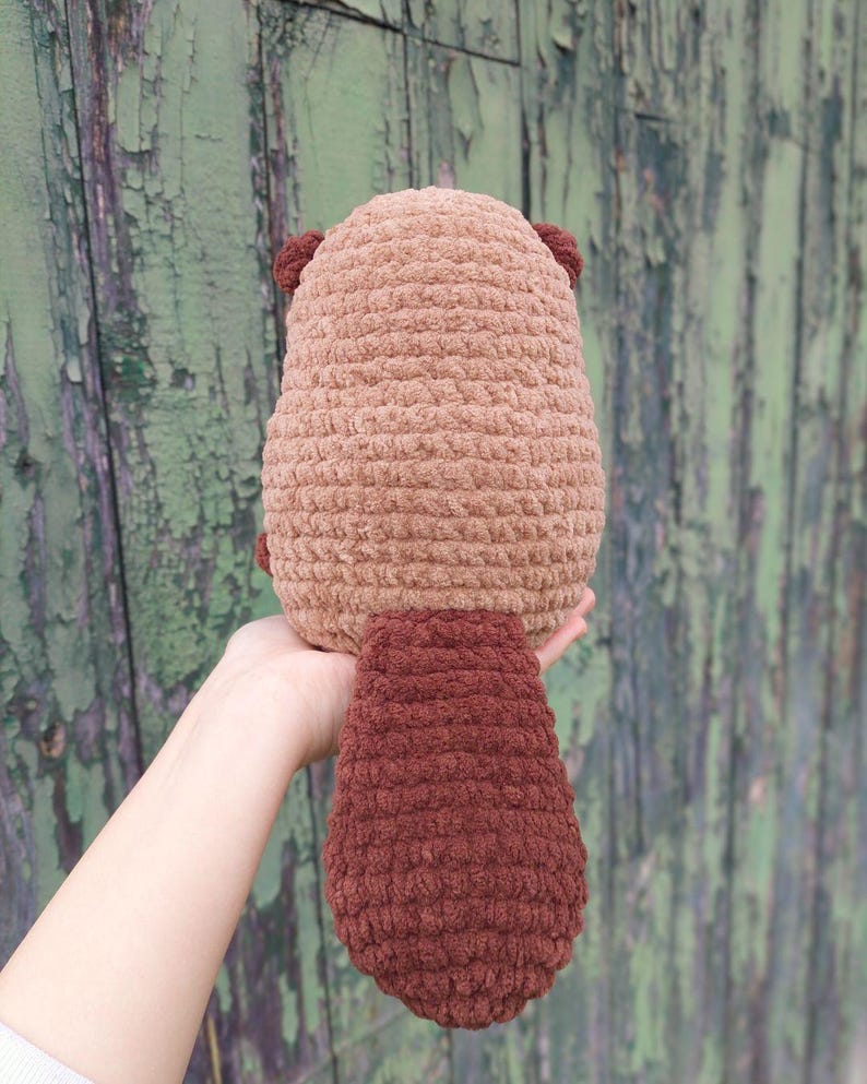 May include: A handmade, plush beaver toy in shades of brown. The body is a light brown, with darker brown ears and a large, flat tail. The toy is made of a soft, textured material, and is designed to be cuddly.