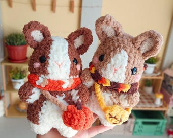 May include: Two plush crochet guinea pigs with soft brown and white yarn. One wears an orange scarf and carries a small orange pumpkin bag, while the other wears a brown and orange scarf with a yellow leaf-shaped bag. Both have black eyes.