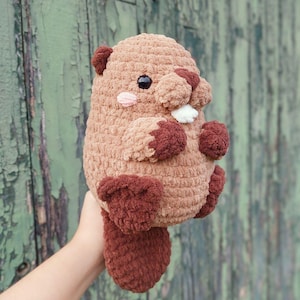 May include: A handmade, plush beaver toy in shades of brown and burgundy. The stuffed animal has a rounded body, small arms and legs, and a flat tail. It features a pink cheek blush, black eyes, and white teeth.