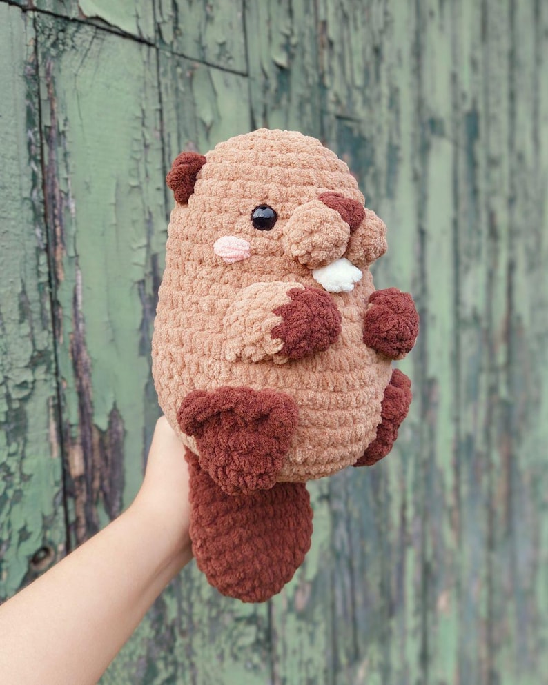 May include: A handmade, plush beaver toy in shades of brown and burgundy. The stuffed animal has a rounded body, small arms and legs, and a flat tail. It features a pink cheek blush, black eyes, and white teeth.