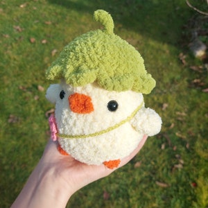 May include: A crocheted stuffed toy chick wearing a green crocheted leaf hat. The chick is white with orange feet and a yellow beak. It has black eyes and a pink flower on its chest.