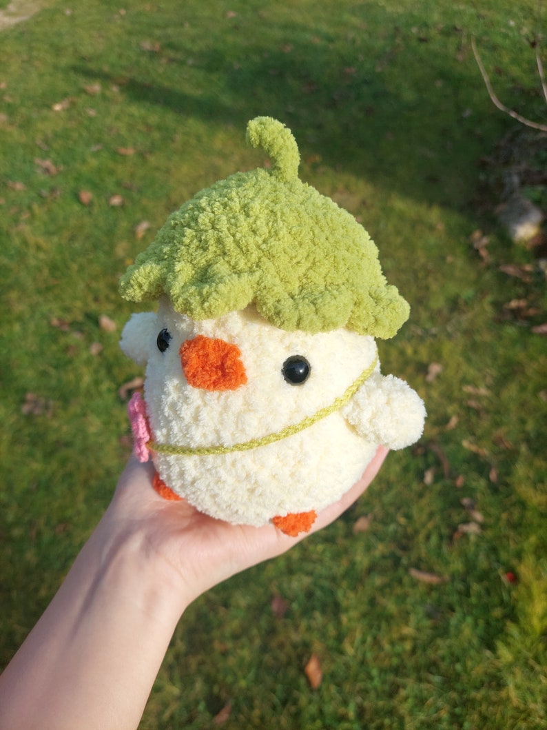 May include: A crocheted stuffed toy chick wearing a green crocheted leaf hat. The chick is white with orange feet and a yellow beak. It has black eyes and a pink flower on its chest.