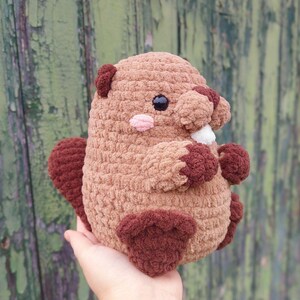 May include: A handmade crochet beaver plushie in shades of brown and tan. The plush toy has dark brown accents on its ears, tail, and feet, with a light pink nose and a white tooth. The beaver has black safety eyes.