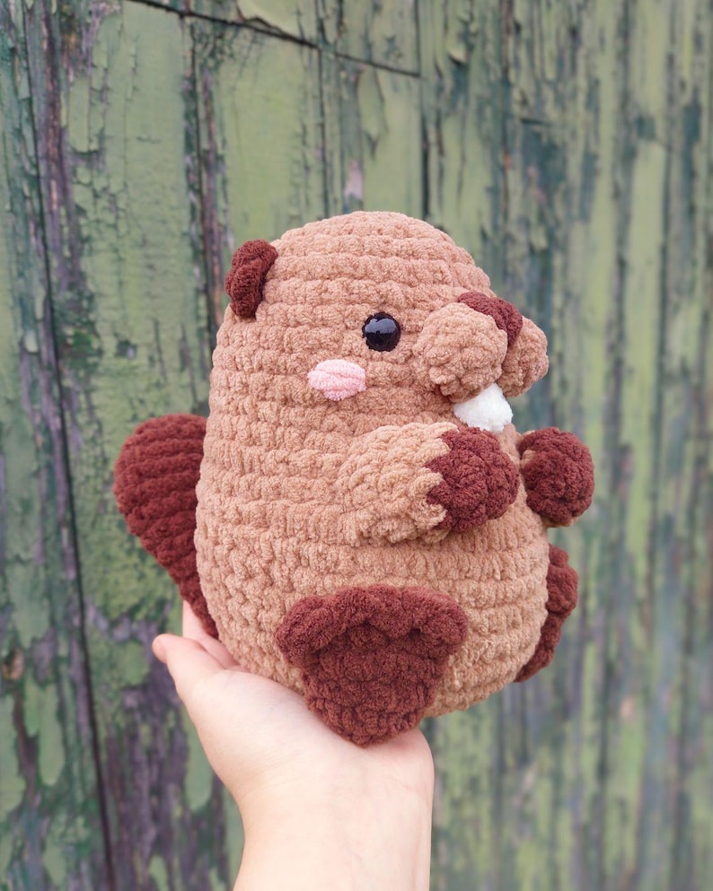 May include: A handmade crochet beaver plushie in shades of brown and tan. The plush toy has dark brown accents on its ears, tail, and feet, with a light pink nose and a white tooth. The beaver has black safety eyes.