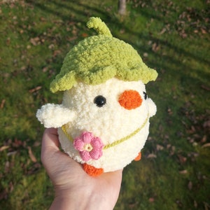 May include: A crocheted chick toy with a green leaf hat, a pink flower, and orange feet. The chick is white with black eyes and an orange beak.