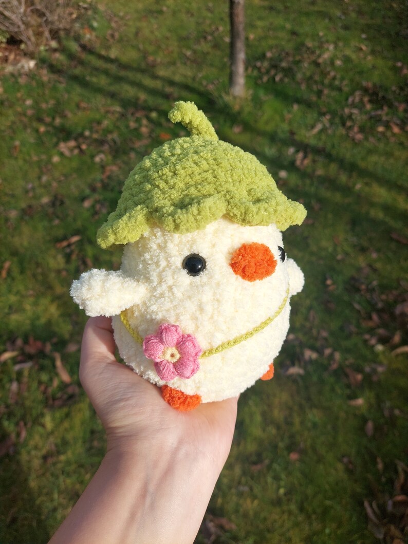 May include: A crocheted chick toy with a green leaf hat, a pink flower, and orange feet. The chick is white with black eyes and an orange beak.