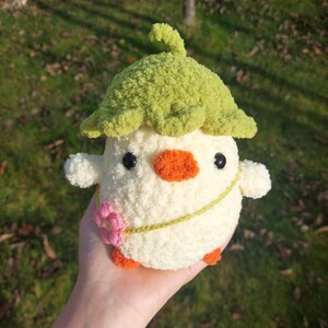 May include: A crocheted stuffed toy chick wearing a green crocheted hat with a flower on its chest. The chick is white with orange feet and a black and orange beak.