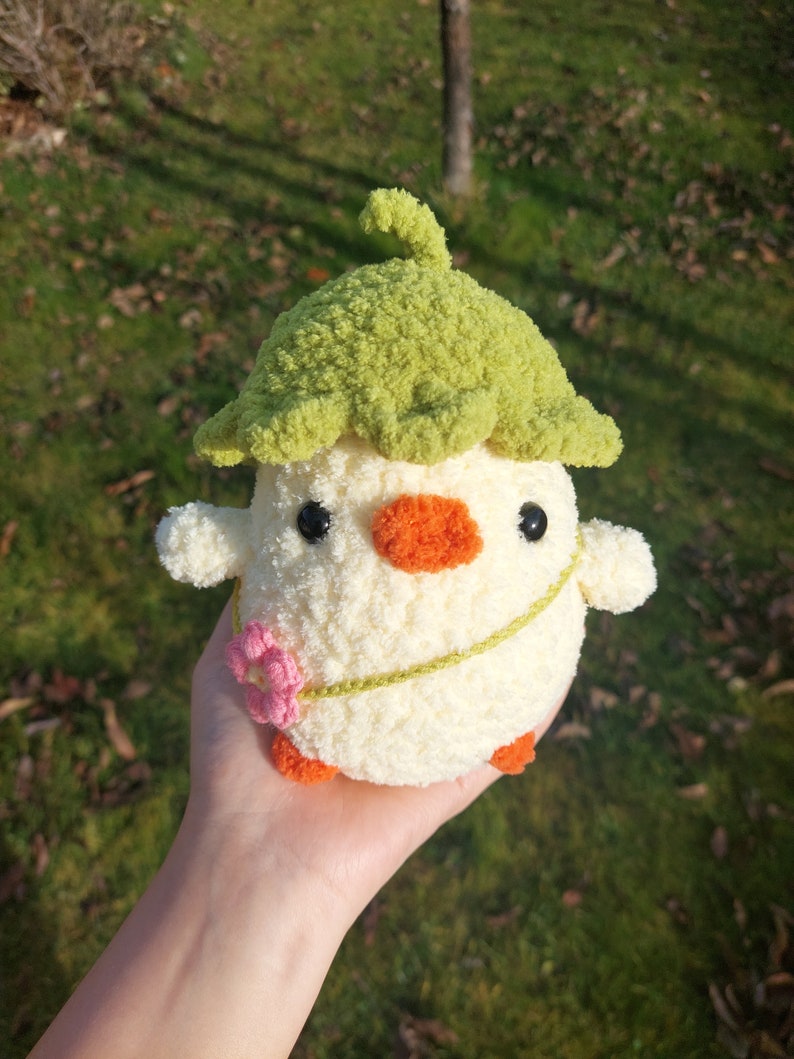 May include: A crocheted stuffed toy chick wearing a green crocheted hat with a flower on its chest. The chick is white with orange feet and a black and orange beak.