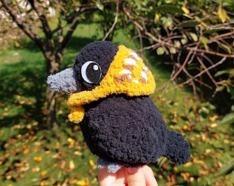 May include: A handmade crochet bird plushie featuring a black textured body, a gray beak, and a yellow hood with white and brown accents. The bird has a large white and black eye and small gray feet, crafted from soft yarn in a rounded shape.