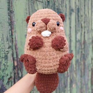 May include: A handmade, plush beaver toy in shades of brown. The beaver has a rounded body, a flat tail, and small, dark brown ears. It features a white tuft of fur and pink cheeks, giving it a cute, friendly appearance. The toy is held against a green background.