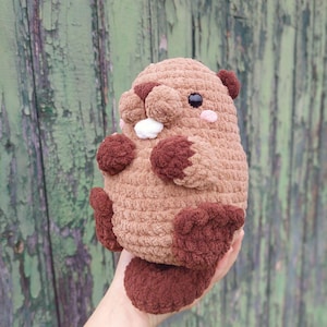 May include: A handmade, plush, brown beaver toy with dark brown accents. The beaver has a round body, small ears, and a fluffy tail. It features black eyes, pink cheeks, and white teeth. The toy is held in a person's hand.