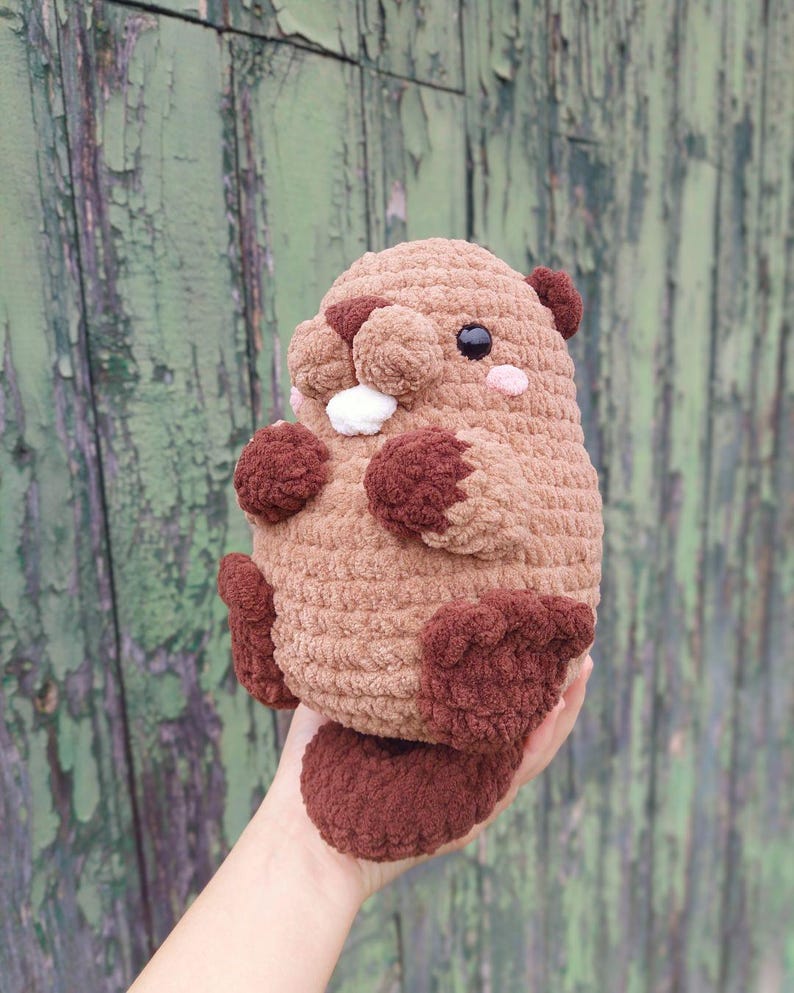 May include: A handmade, plush, brown beaver toy with dark brown accents. The beaver has a round body, small ears, and a fluffy tail. It features black eyes, pink cheeks, and white teeth. The toy is held in a person's hand.