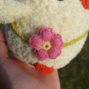 May include: A crocheted plush toy chick with a pink flower on its tummy. The chick is yellow with a green leaf on its head and orange feet.