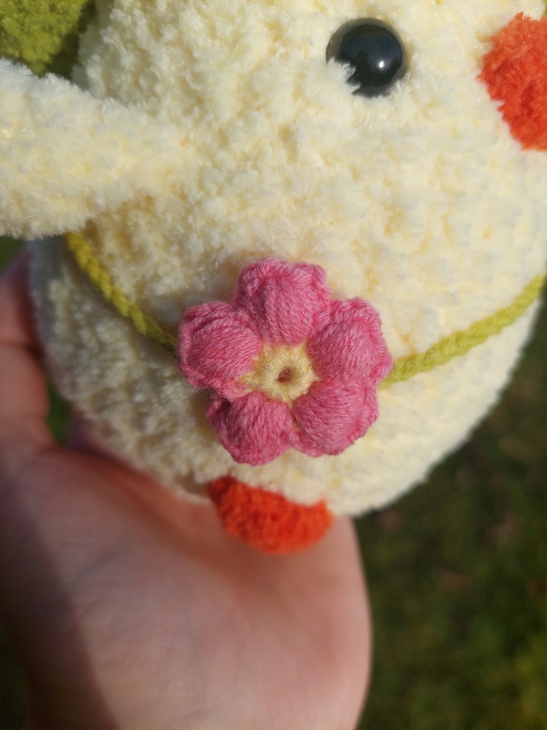 May include: A crocheted plush toy chick with a pink flower on its tummy. The chick is yellow with a green leaf on its head and orange feet.