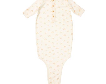 May include: Cream-colored baby sleep sack with a pattern of small, peach-colored rainbows. The sleep sack has long sleeves, a button-up front, and wooden buttons. The fabric appears soft and comfortable, suitable for a baby.