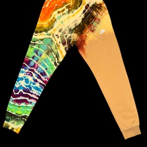 Pode incluir: Calças de moletom tie-dye com um design dividido. Uma perna apresenta um padrão vibrante e multicolorido de laranja, amarelo, verde, azul e roxo. A outra perna é bege sólido com um pequeno logotipo branco da Nike. As calças têm cintura com cordão e punhos canelados.