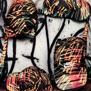 May include: Four black baseball caps with a colourful abstract paint splatter design. The caps have a curved brim and adjustable strap. One cap features a white Nike logo.