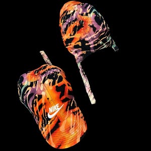 May include: Two Nike baseball caps with a vibrant abstract pattern in orange, purple, and black. The caps have a curved peak and an adjustable strap. The Nike logo is visible on the front of one cap.