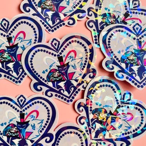 May include: Heart-shaped stickers with a holographic finish. Each sticker features a cartoon illustration of two characters in a loving embrace, set against a light gray background with a blue border. The stickers are arranged on a pink surface.