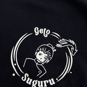 Op de afbeelding: Zwart T-shirt met een wit grafisch ontwerp van een cartoonfiguur met de tekst "Geto Suguru".