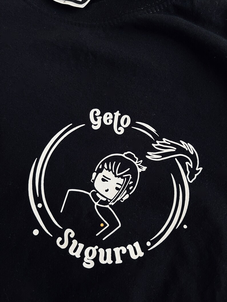 Op de afbeelding: Zwart T-shirt met een wit grafisch ontwerp van een cartoonfiguur met de tekst "Geto Suguru".
