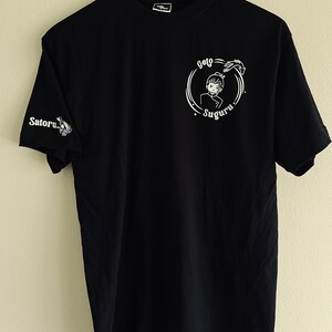 Op de afbeelding: Zwart T-shirt met witte tekst en een afbeelding van een personage in een cirkel. De tekst luidt "Geto Suguru".