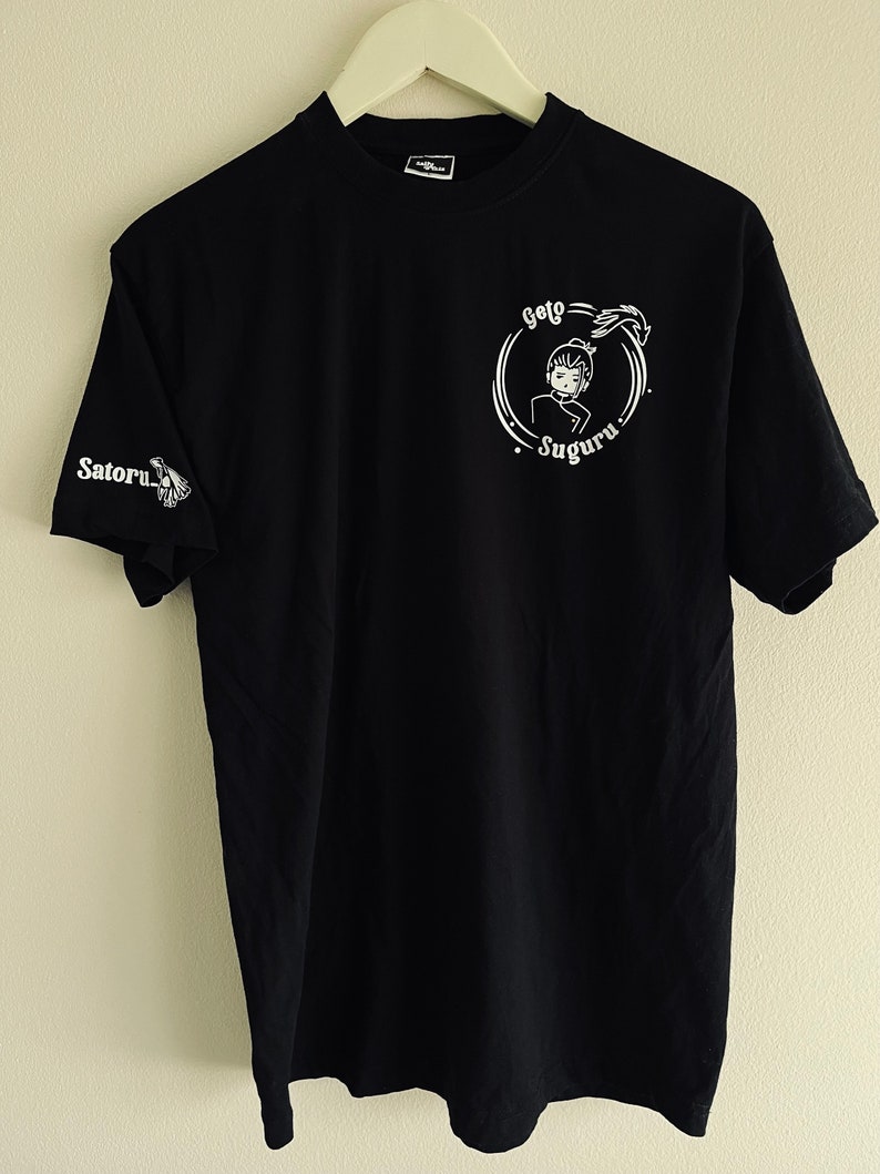 Op de afbeelding: Zwart T-shirt met witte tekst en een afbeelding van een personage in een cirkel. De tekst luidt "Geto Suguru".