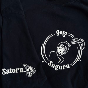 Op de afbeelding: Zwart T-shirt met een witte afbeelding van een personage met de tekst "Geto Suguru" en "Satoru" op de mouw.