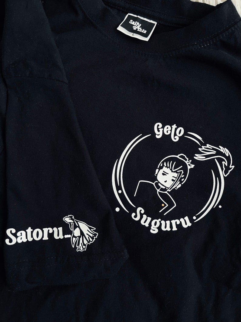 Op de afbeelding: Zwart T-shirt met een witte afbeelding van een personage met de tekst "Geto Suguru" en "Satoru" op de mouw.
