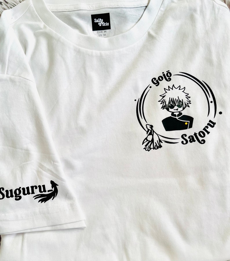 Op de afbeelding: Wit T-shirt met een zwart-witte grafische print van een personage met de tekst "Gojo Satoru" en een kleinere grafische print op de mouw die "Suguru" zegt.