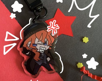 Puede incluir: Un llavero de personaje de anime rojo, blanco y negro con el texto "CHUUYA!" sobre un fondo rojo. El personaje lleva un sombrero negro y una chaqueta negra. El llavero está unido a un anillo de metal negro.