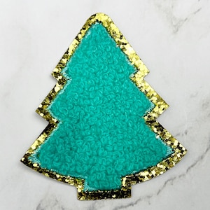 May include: A teal Christmas tree-shaped patch with a gold glitter border. The tree has a textured, looped surface. The patch is set against a white and gray marble background.